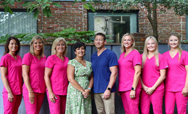 A group of women wearing scrubs pose together with a man, likely for a team photo, set against an outdoor backdrop.