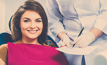 A smiling woman wearing a dental hygienist s uniform and sitting in a dentist s chair with a dental professional behind her, discussing paperwork.