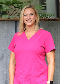 The image shows a woman wearing a pink scrub top, standing outdoors with a smile, posing for a portrait.