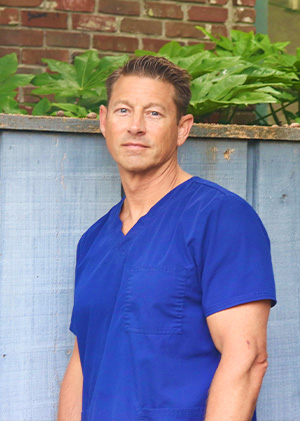 The image shows a man standing against a fence, dressed in blue scrubs, with dark hair and a beard, wearing glasses, and looking directly at the camera.