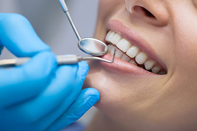 The image shows a person receiving dental care with a dental hygienist using a tool to clean their teeth while wearing a blue glove, with the patient smiling and appearing content.