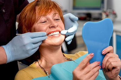 A woman sitting in a dental chair with a smile on her face, receiving dental care from a professional who is adjusting her teeth and wearing a blue mask over her mouth.