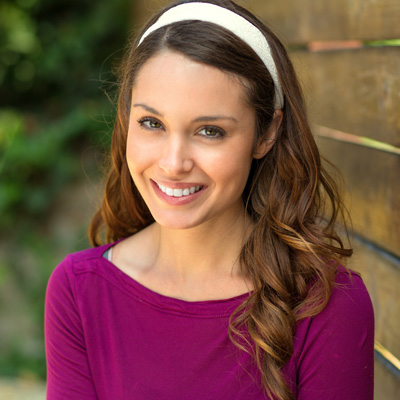 A smiling woman with long hair and a purple top poses against a wooden fence.