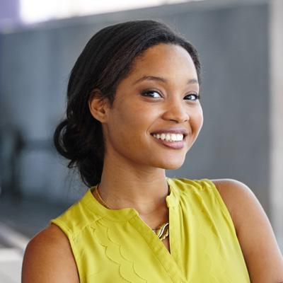 A smiling woman with dark hair, wearing a yellow top, stands against a blurred background.