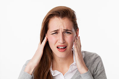 The image shows a woman with her hands on her temples, appearing to be experiencing discomfort or pain. She has a concerned expression and is wearing a light-colored top.