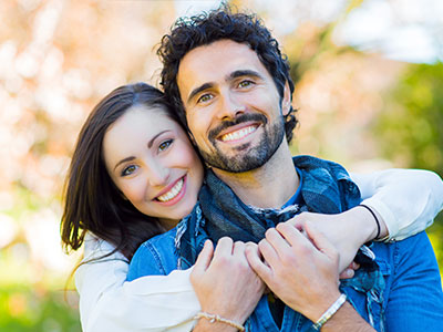 The image shows a man and woman embracing each other outdoors with smiles on their faces  they are both wearing casual attire and appear to be enjoying a warm moment together.