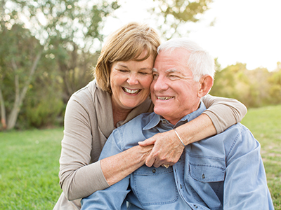 The image depicts an elderly man and woman embracing each other outdoors during daylight.