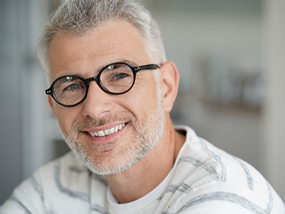 The image shows a man with grey hair and glasses, smiling at the camera while looking directly into it. He has facial stubble and appears to be middle-aged.