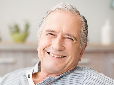 The image shows an older man with gray hair smiling at the camera while seated comfortably indoors.