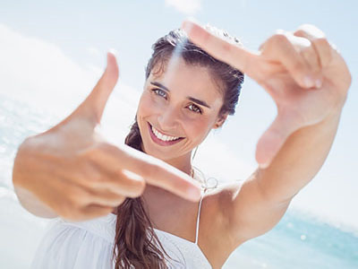 A smiling woman with long hair holds up her hand to frame a picture of herself against a bright blue sky background.